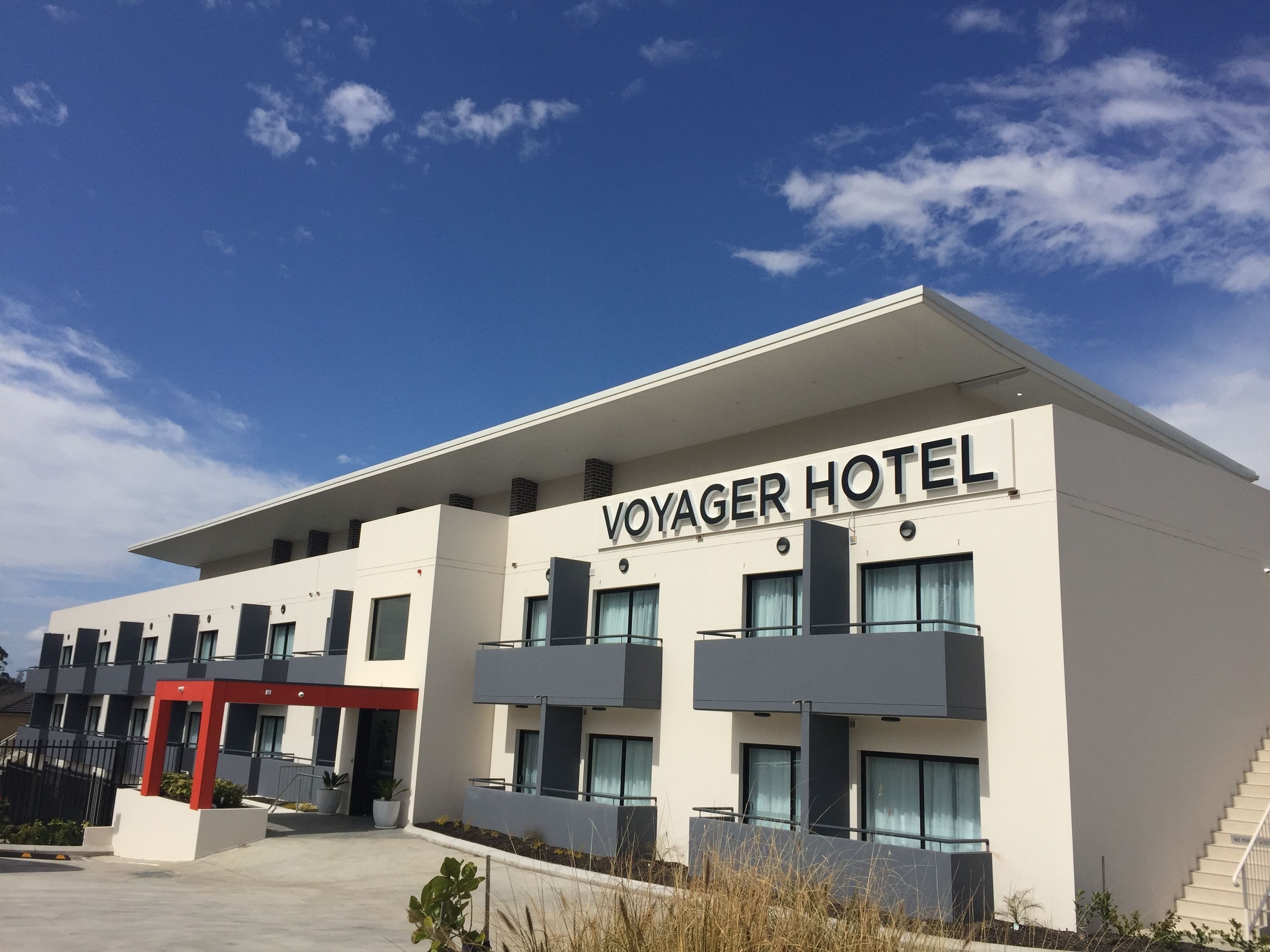Un bâtiment moderne avec l’enseigne « VOYAGER HOTEL » sur la façade, caractérisé par un toit plat, de grandes fenêtres et une structure d’accentuation de couleur rouge.