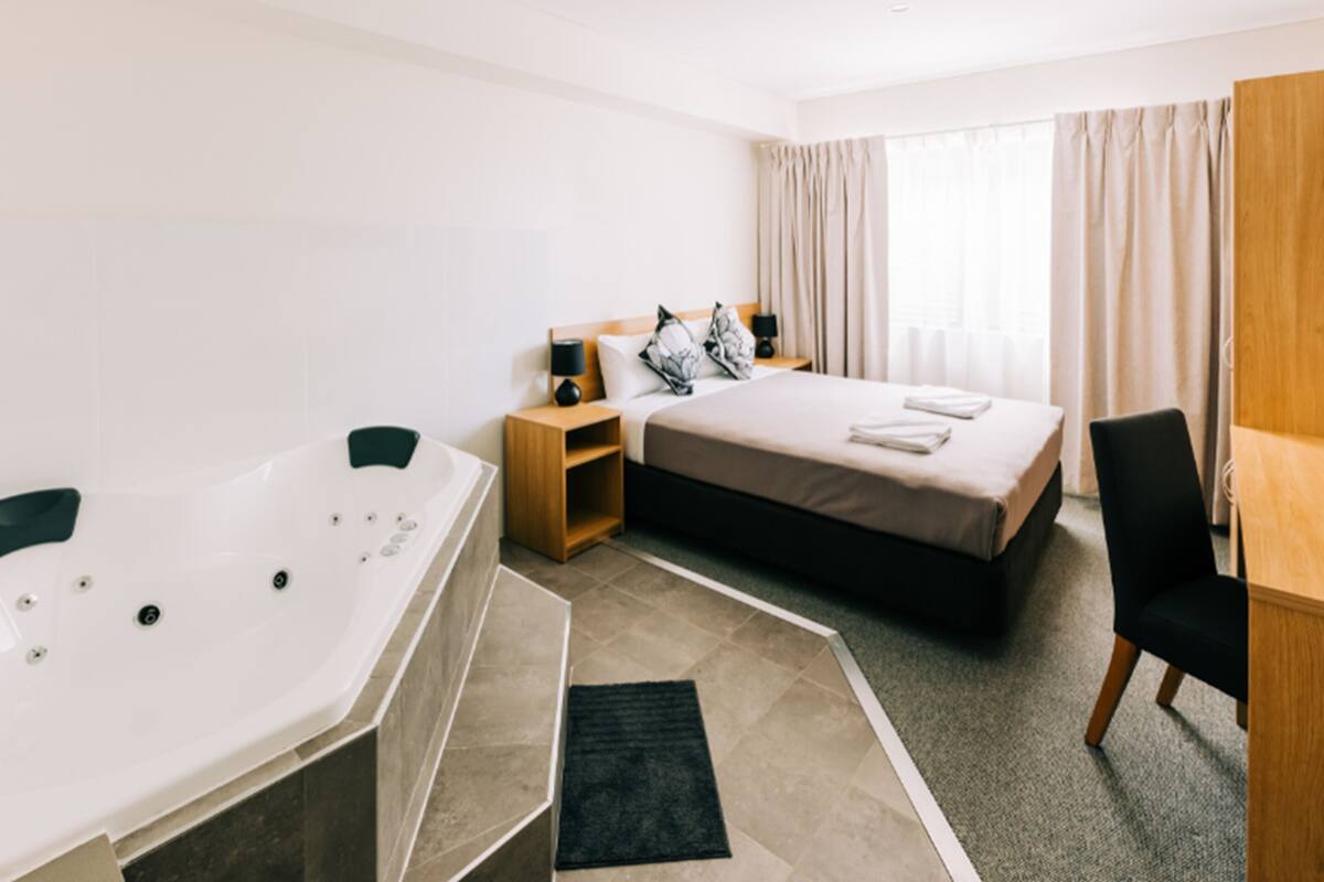 A hotel room with a single bed, a bedside table with a lamp, a small wooden nightstand, a black chair, a desk, and a large white bathtub.