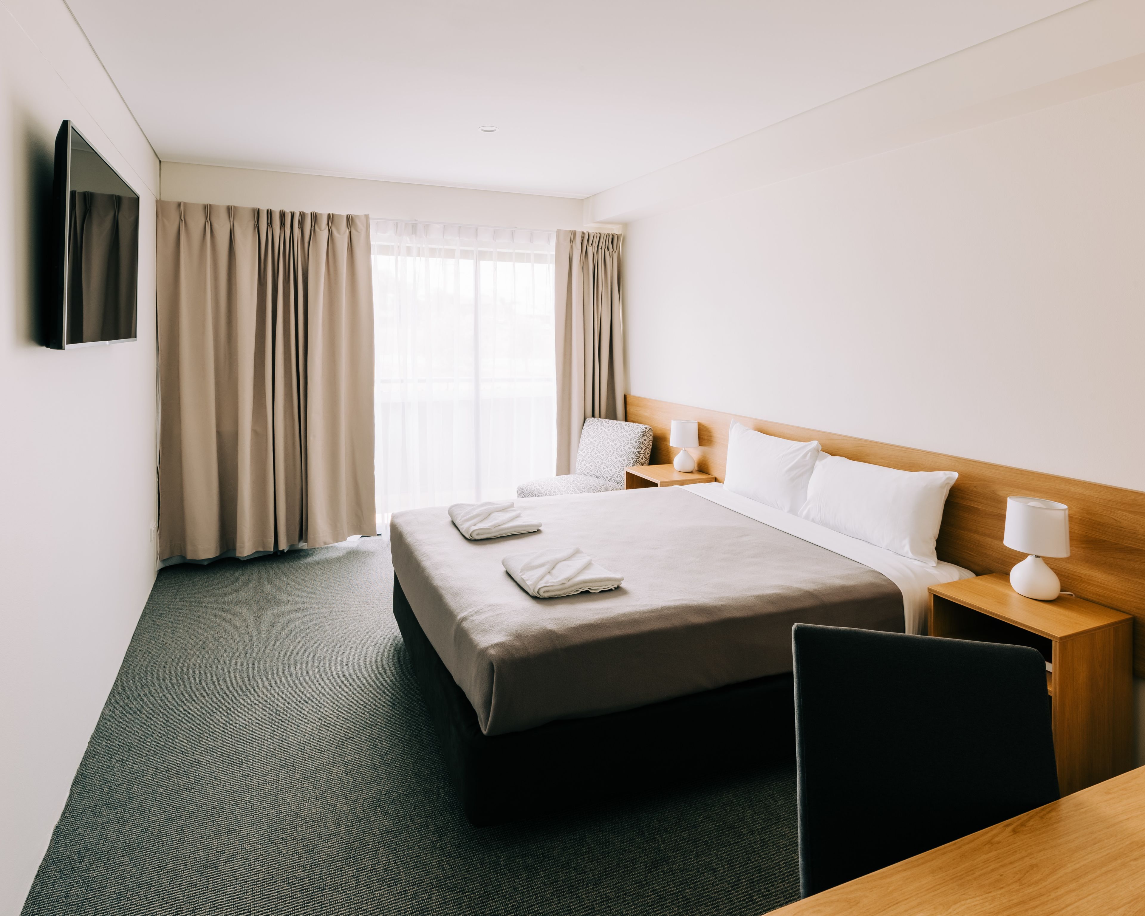 Quarto de hotel com cama, escrivaninha, cadeira, TV fixada na parede e janela com cortinas.
