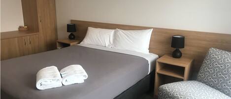 A hotel room with a bed, two bedside tables with lamps, a wooden wardrobe, and a patterned armchair.