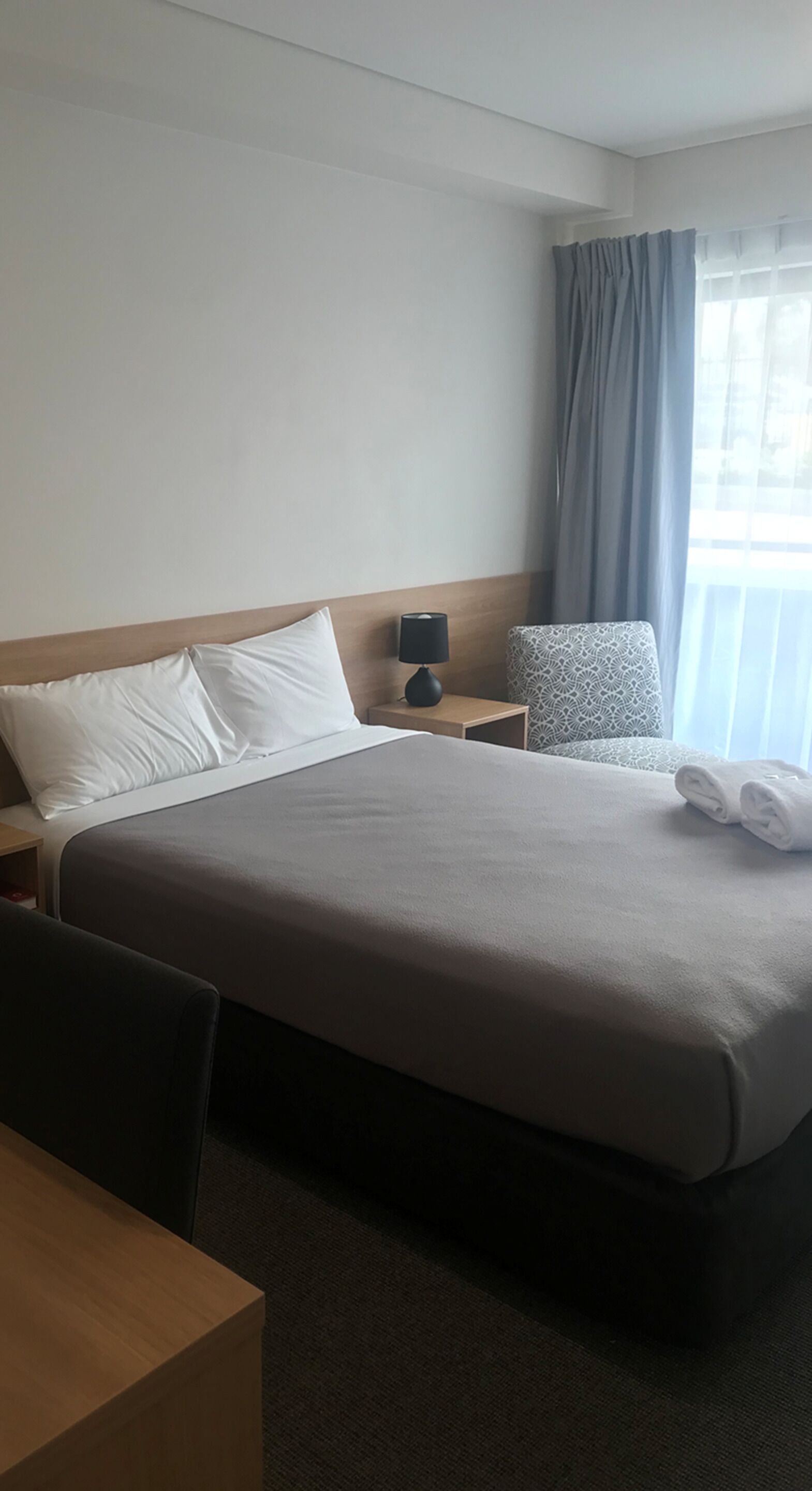 Ett hotellrum med en stor säng, ett nattduksbord med en lampa, en stol och ett fönster med gardiner.
