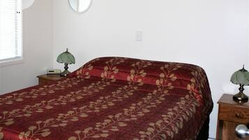 Un dormitorio con una cama grande, una mesita de noche con lámpara, una luz en la pared, una ventana con persianas y un espejo.