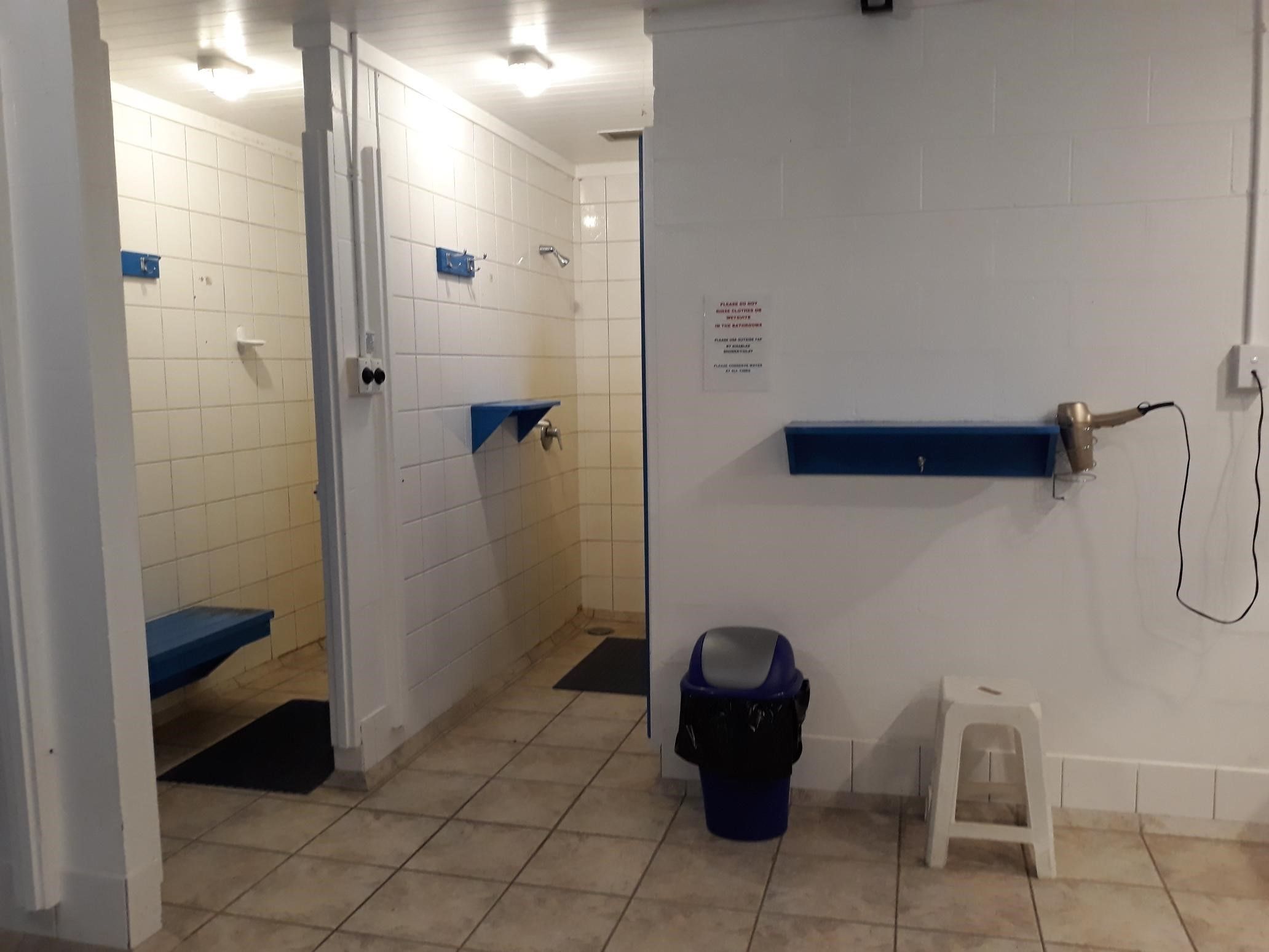 Un baño público con paredes de azulejos blancos, bancos azules, un cubo de basura y un taburete pequeño.
