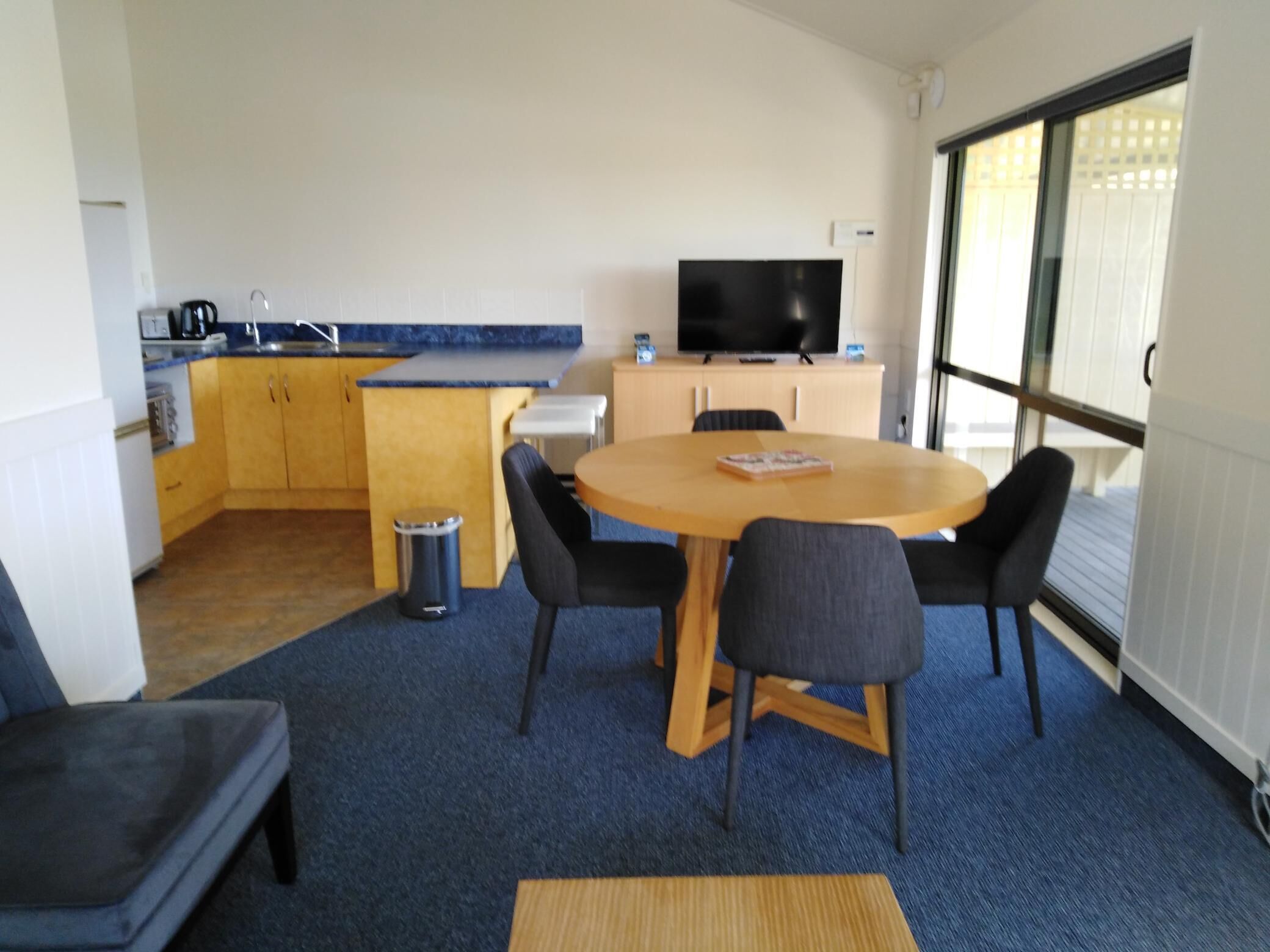 A compact room with a round wooden table, four chairs, a small kitchen area with a sink and cabinets, a television on a stand, and a large window providing natural light.