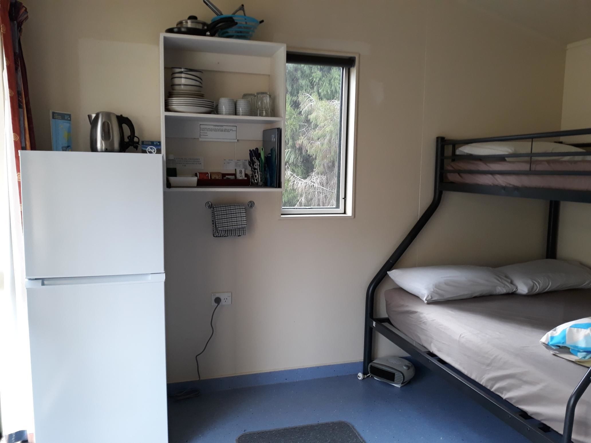A compact room with a bunk bed, a small kitchenette with a kettle and dishes, and a window with a view of trees.