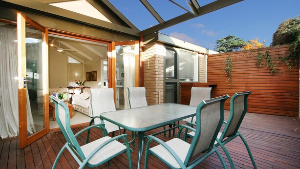 A patio with a table and chairs, a glass door leading to a room, and a wooden fence with plants. - Wattle Holiday Rental (Hawthorn)