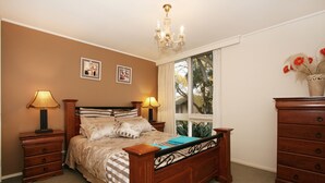 A bedroom with a wooden bed, two bedside tables with lamps, a dresser, and a window with a view of trees. - Wattle Holiday Rental (Hawthorn)