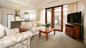 A living room with a sofa, coffee table, and a TV on a wooden cabinet. There is a kitchen area with a refrigerator and microwave in the background. - Wattle Holiday Rental (Hawthorn)
