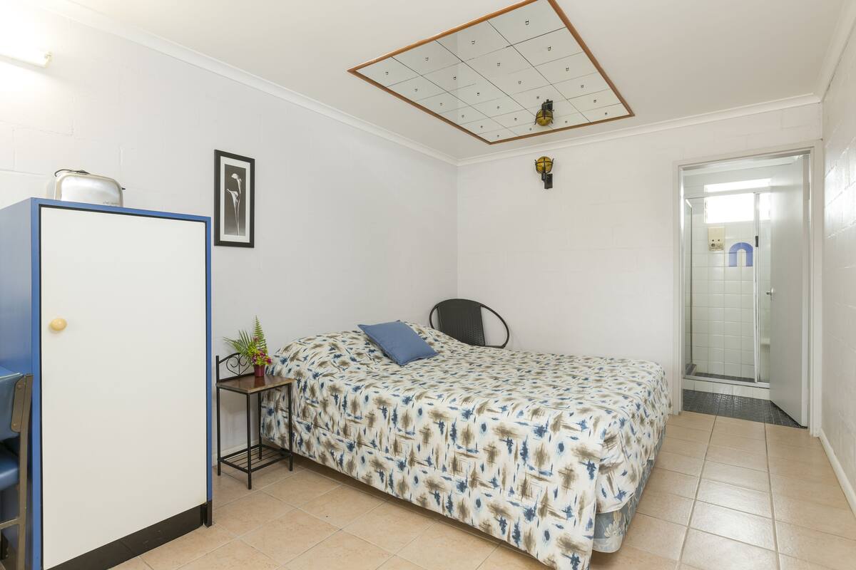 A small, clean room with a bed, a desk, a chair, a wardrobe, and a shower area.