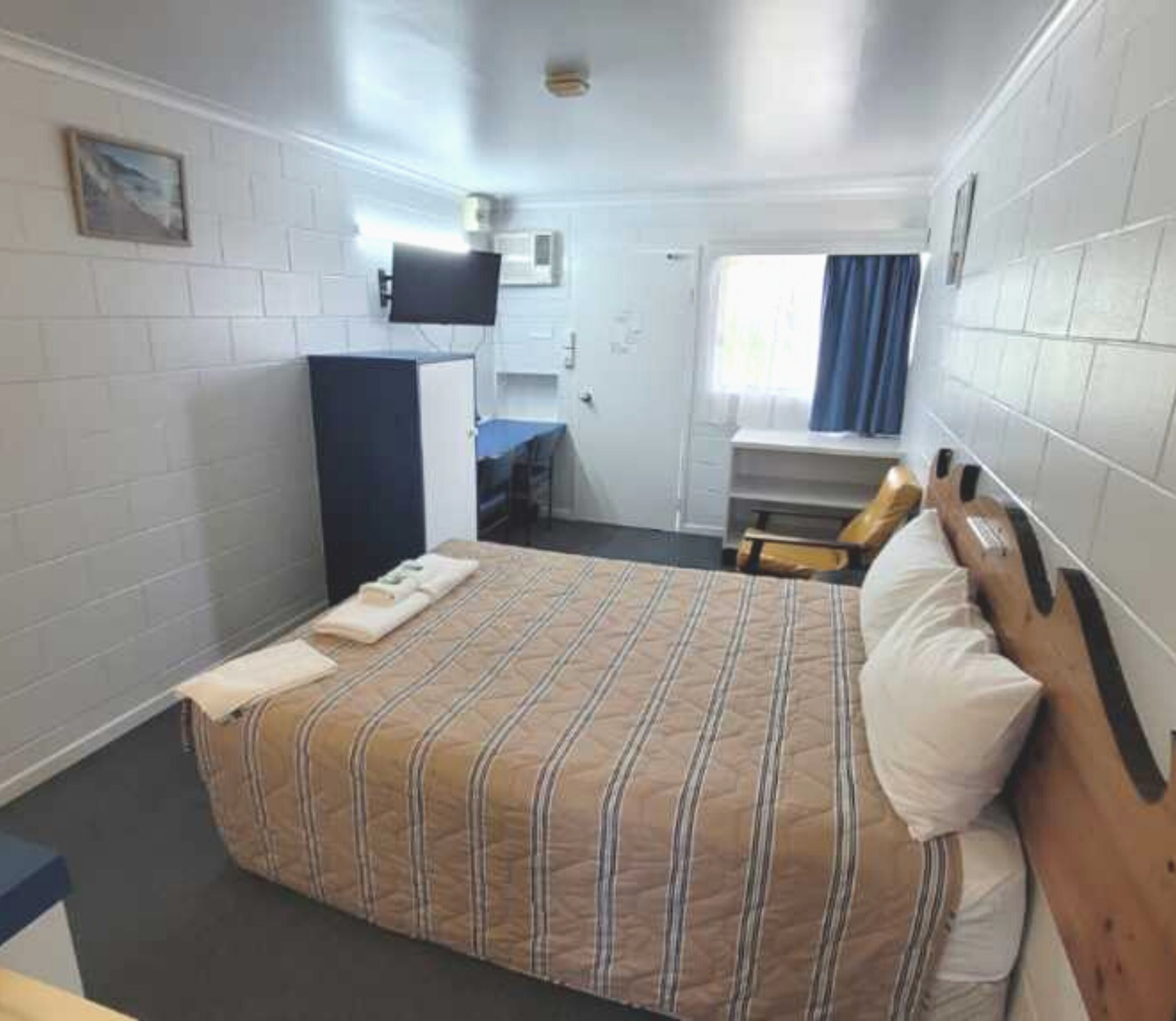 A compact hotel room with a single bed, a desk with a chair, a television mounted on the wall, and a window with curtains.