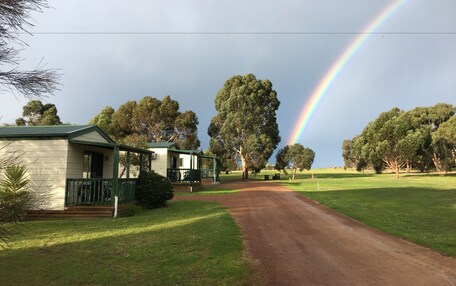 A rural road leading to a row of single-story houses with green railings, surrounded by trees and a visible rainbow.