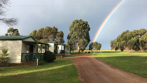 A rural road leading to a row of single-story houses with green railings, surrounded by trees and a visible rainbow. - Kangaroo Island Cabins (Kingscote)