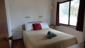 A hotel room with a bed, a window with curtains, a bedside lamp, and a wall-mounted light. - Kangaroo Island Cabins (Kingscote)