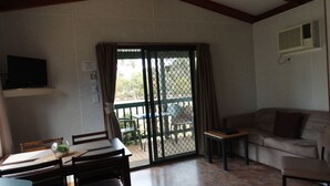 A room with a dining table, chairs, a sofa, a TV mounted on the wall, and a sliding glass door leading to an outdoor area with a table and chairs. - Kangaroo Island Cabins (Kingscote)