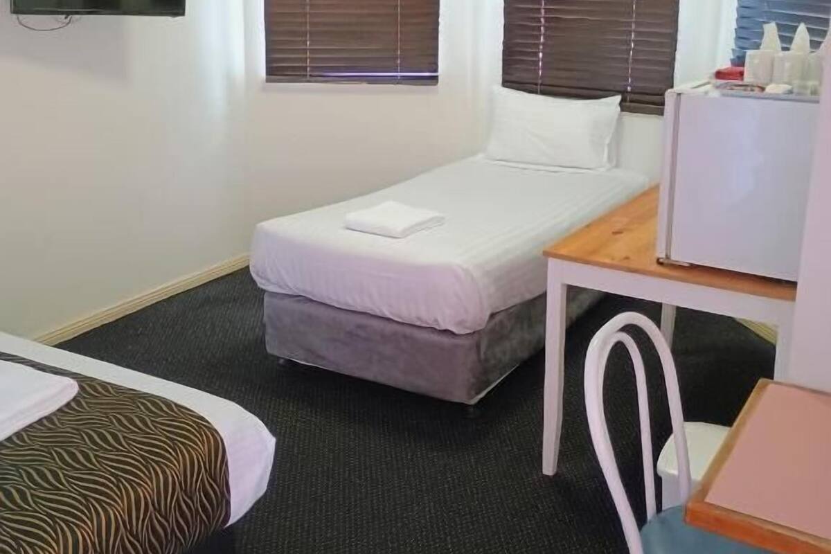 Ein kleines Hotelzimmer mit einem Einzelbett, einem Schreibtisch, einem Stuhl, einem Fernseher und zwei Fenstern mit Jalousien.