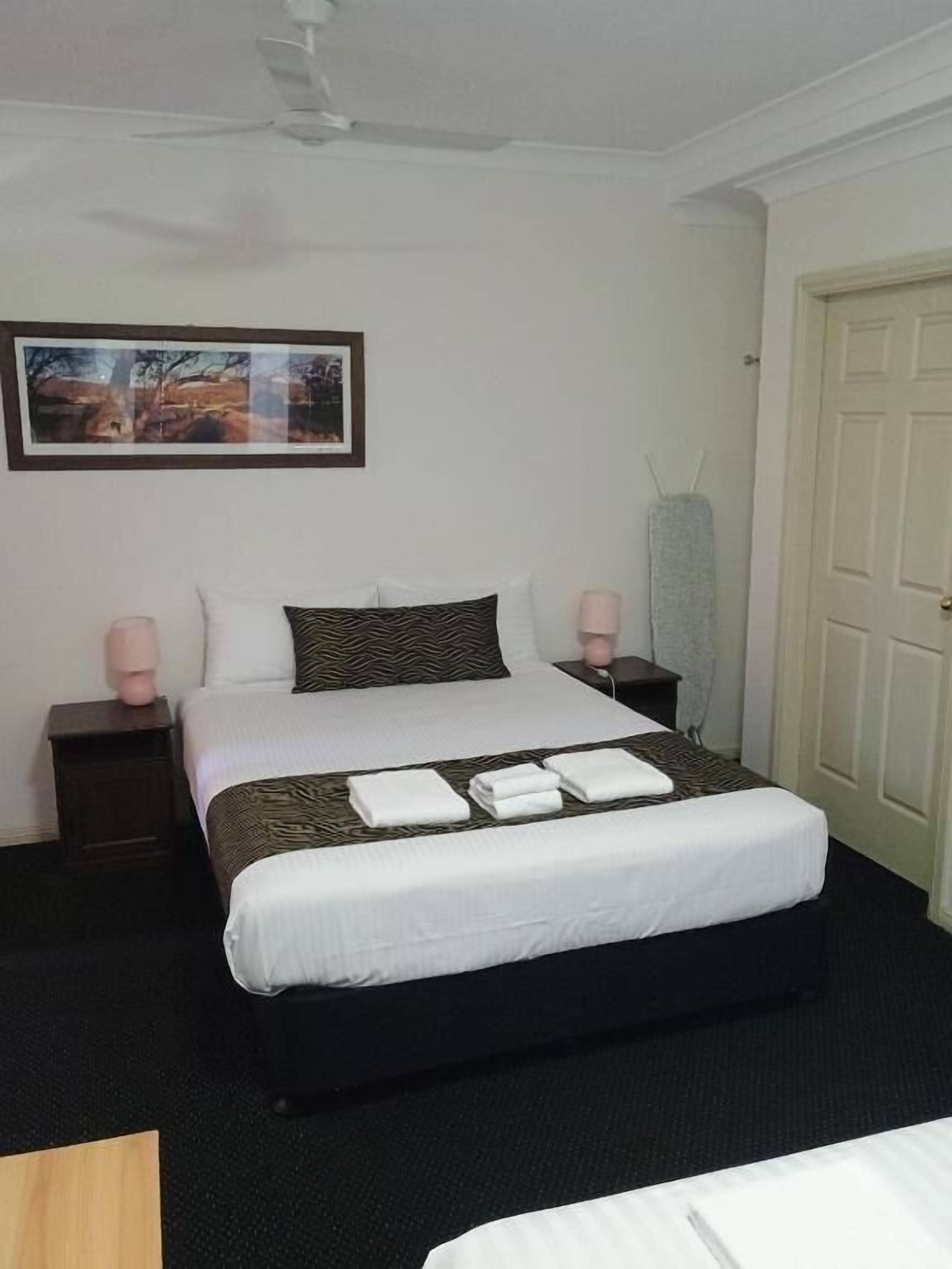 A neatly made bed with white linens and a patterned pillow, two bedside tables with lamps, a framed picture on the wall, and a door to the right.