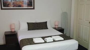 A neatly made bed with white linens and a patterned pillow, two bedside tables with lamps, a framed picture on the wall, and a door to the right.