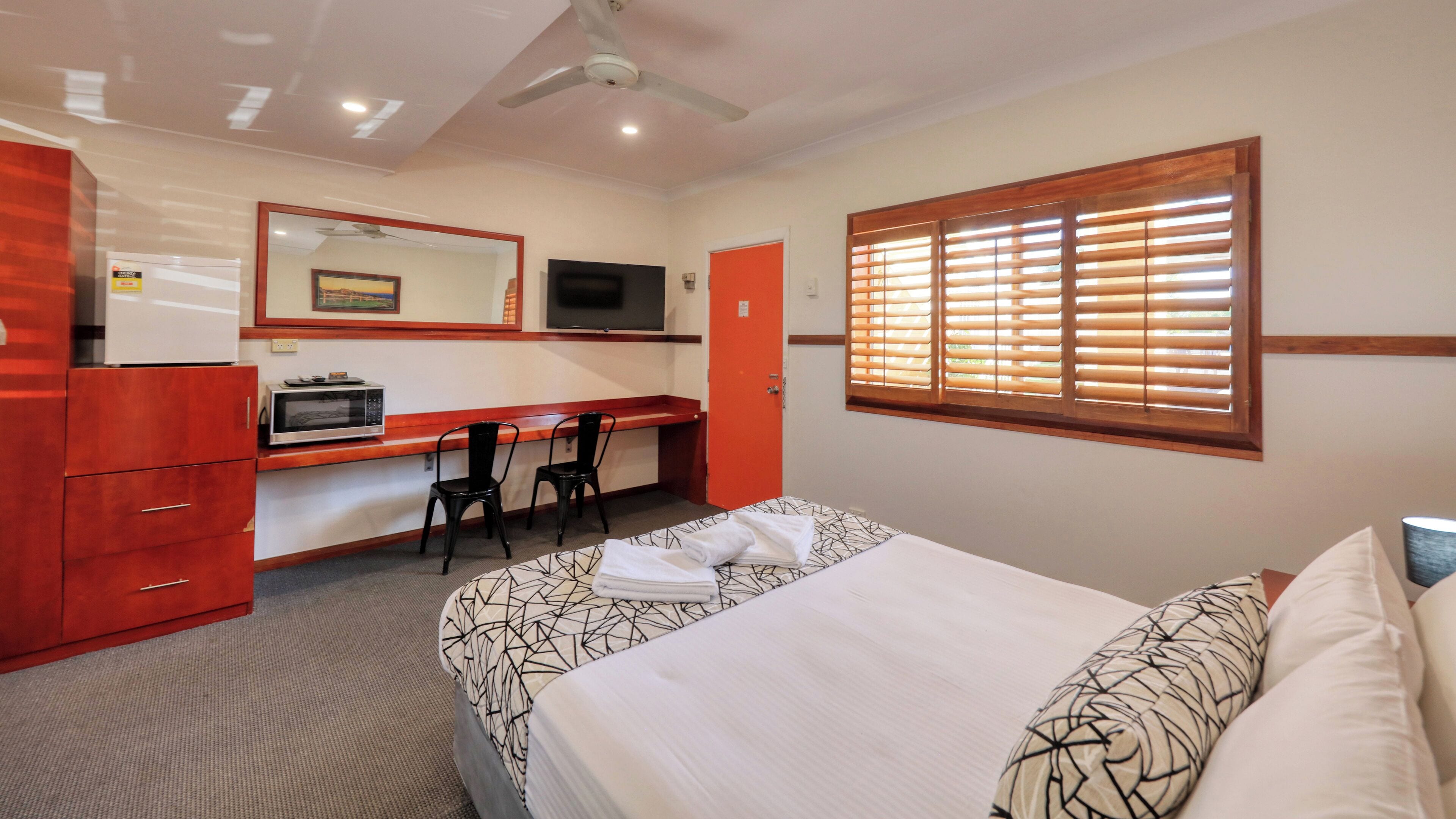 A room with a bed, a desk, a chair, a television, and a window with blinds.