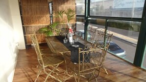 A dining area with a metal table and chairs, a woven wall, and a view of the street through large windows.