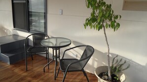 A small outdoor seating area with a table and two chairs, a potted tree, and a window with blinds.