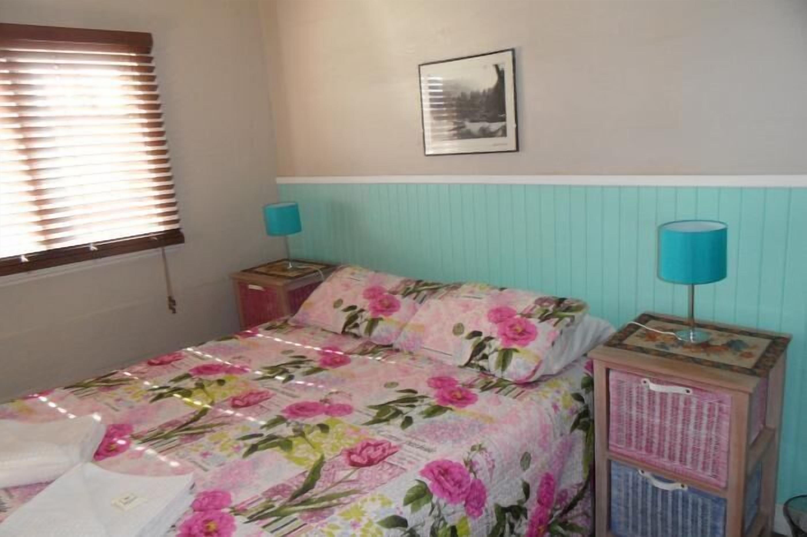 A bedroom with a floral bedspread, a nightstand with a lamp, a framed picture on the wall, and a window with blinds.