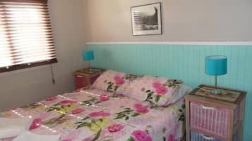 A bedroom with a floral bedspread, a nightstand with a lamp, a framed picture on the wall, and a window with blinds.