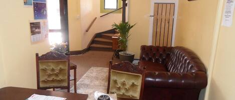 A living room with a brown leather sofa, a dining table with papers, a potted plant, and a staircase in the background.