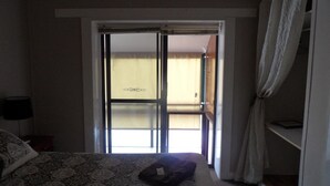 A bedroom with a bed, a nightstand, a lamp, a sliding glass door leading to a balcony, and a wardrobe.