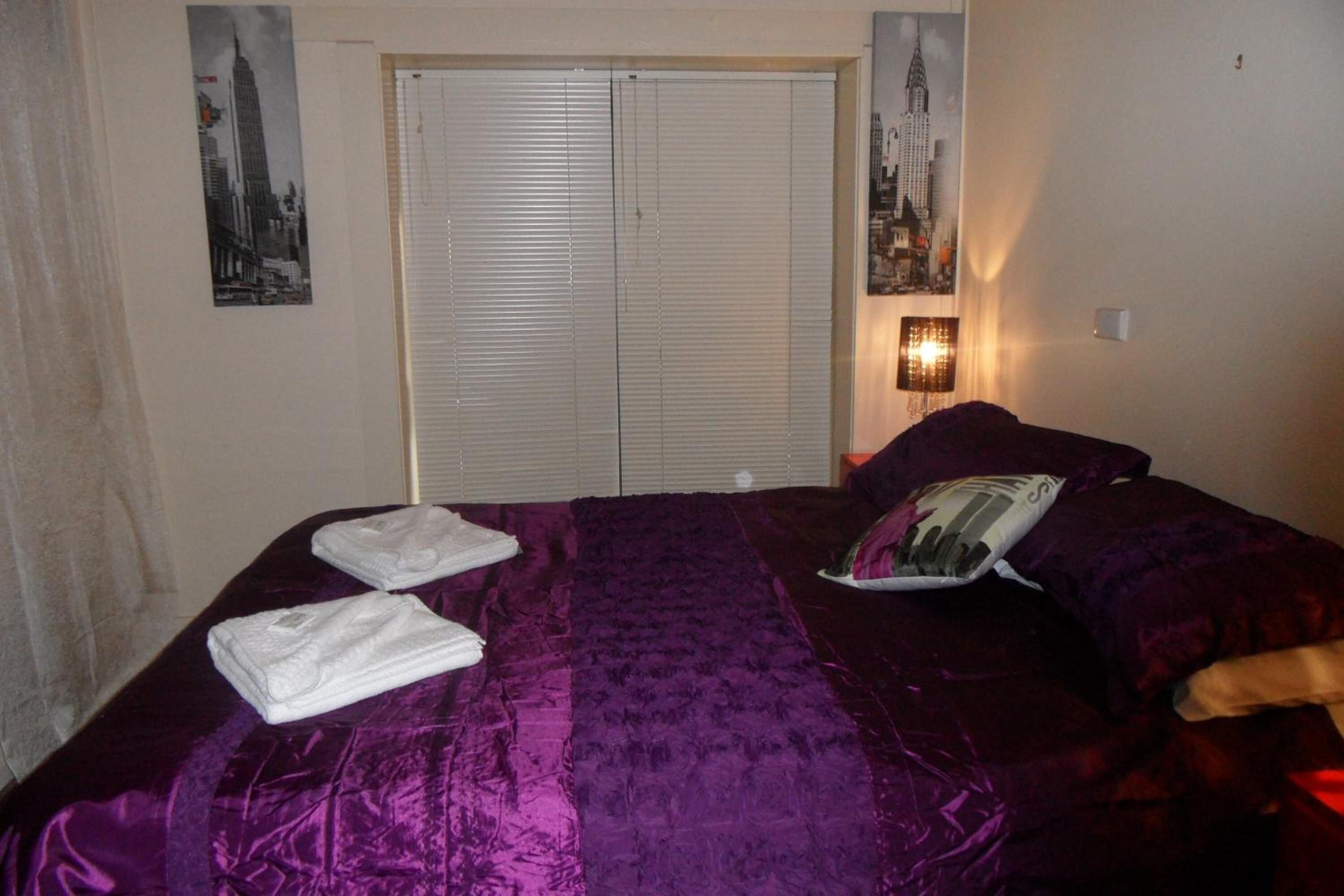 A hotel room with a bed, a door with blinds, and a wall-mounted lamp.