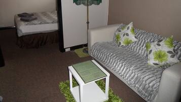 A small, furnished room with a white sofa, a green lamp, a small table, and a bed with a patterned bedspread.