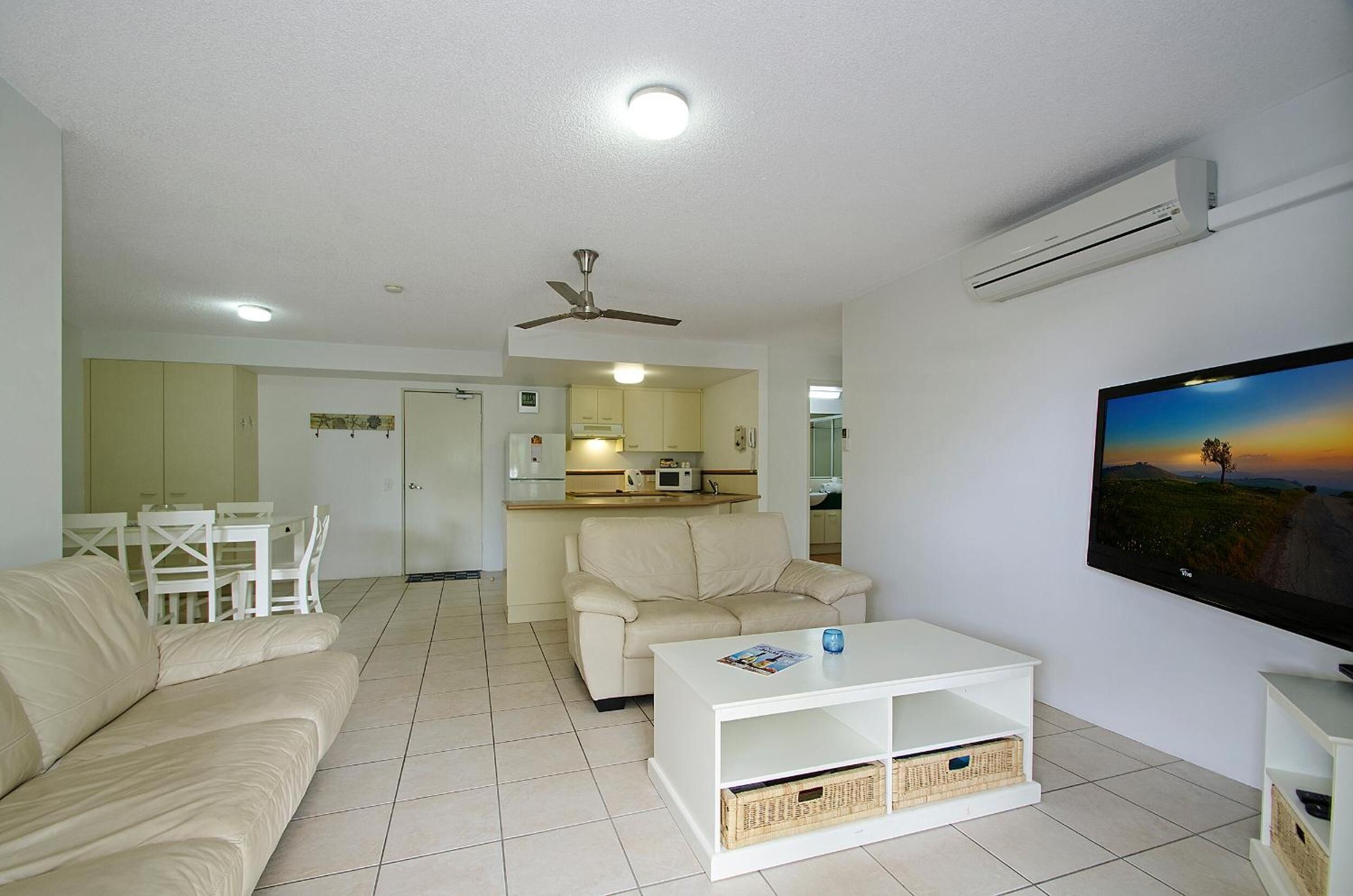 A living room with a sofa, coffee table, TV, and a kitchen area in the background.