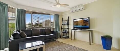Una sala de estar con un sofá negro, una mesa de centro, un televisor sobre un mueble bajo, un ventilador de techo y un ventanal con cortinas.