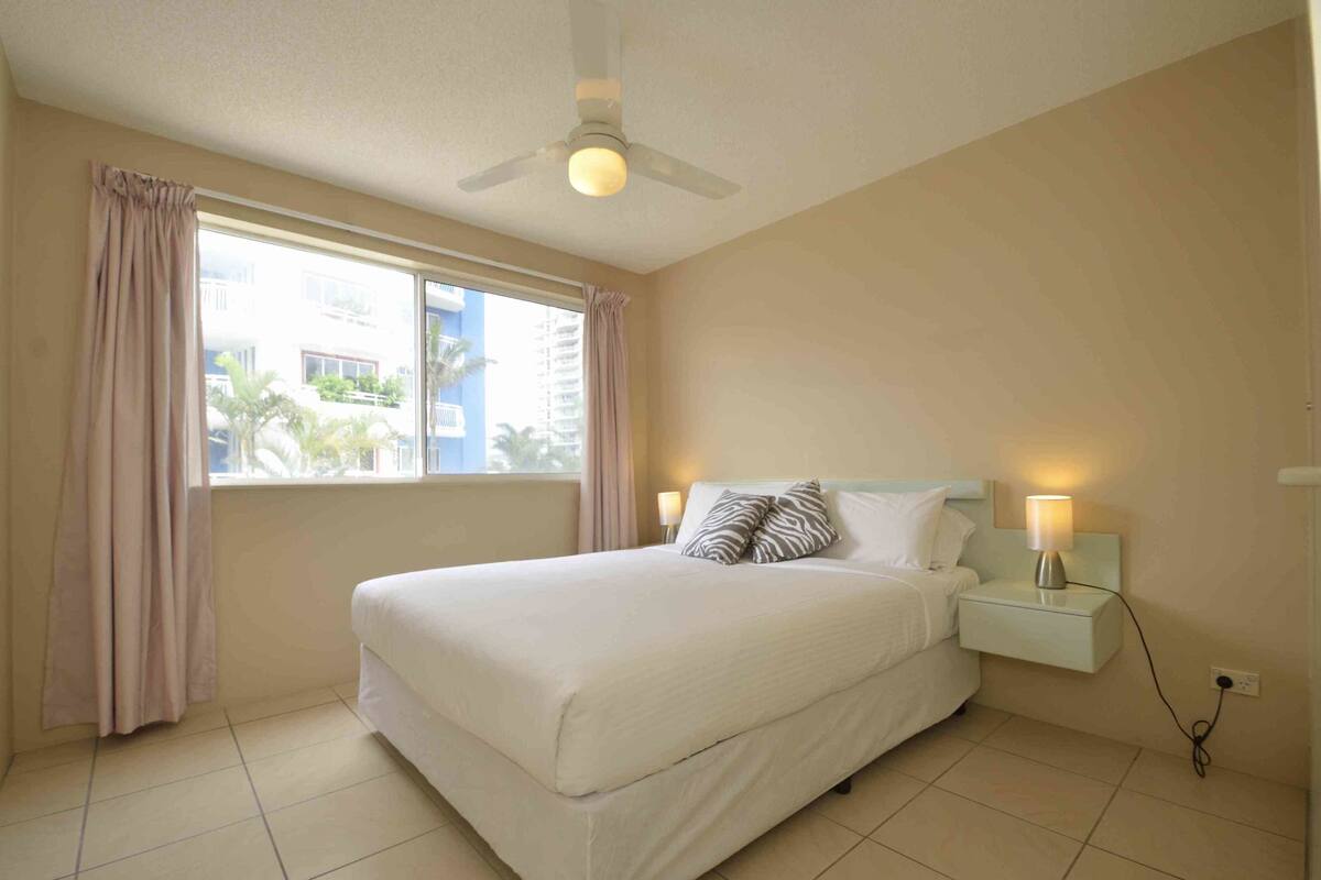 A hotel room with a bed, a nightstand, a lamp, a ceiling fan, and a window with curtains.