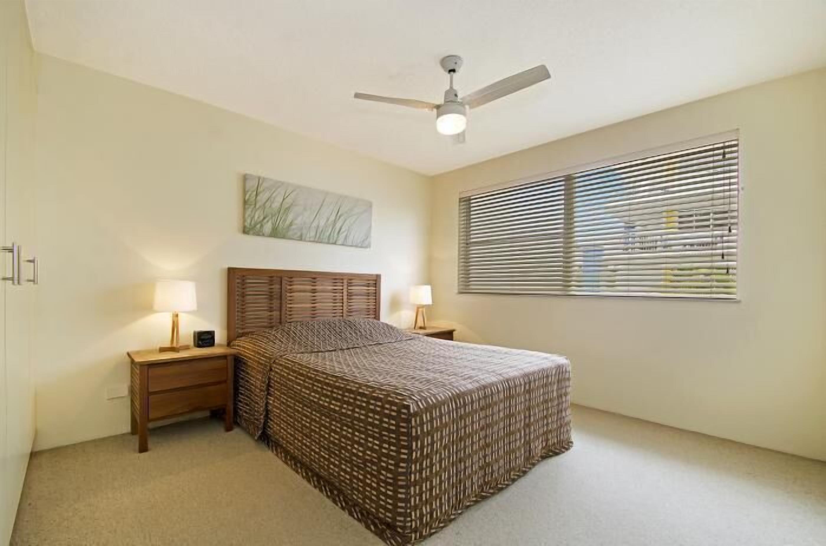 Un dormitorio con cama, dos mesitas de noche con lámparas, ventilador de techo, ventana con persianas y una obra de arte colgada en la pared.
