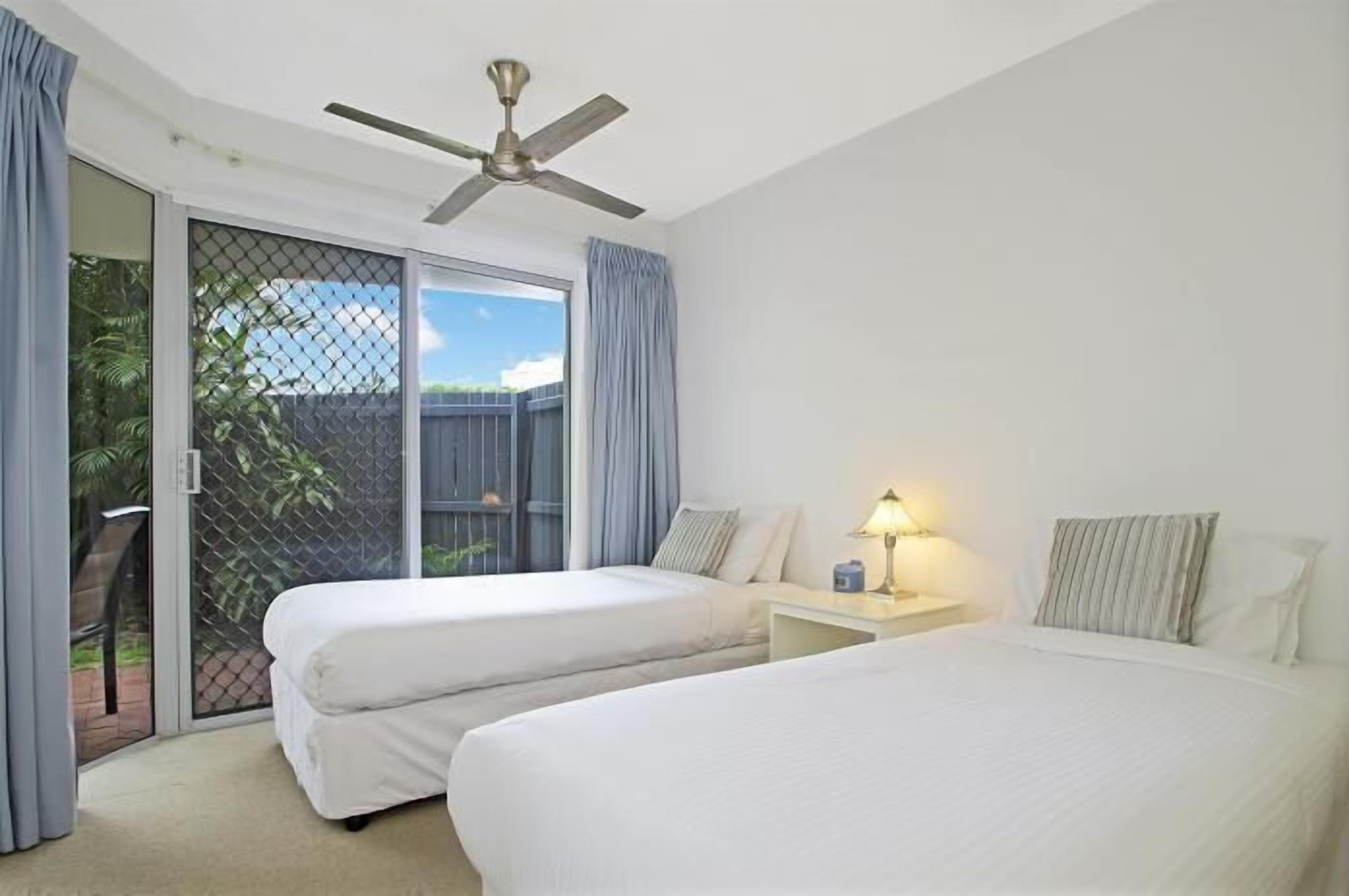 A hotel room with two beds, a ceiling fan, a nightstand with a lamp, and a sliding glass door leading to an outdoor area.