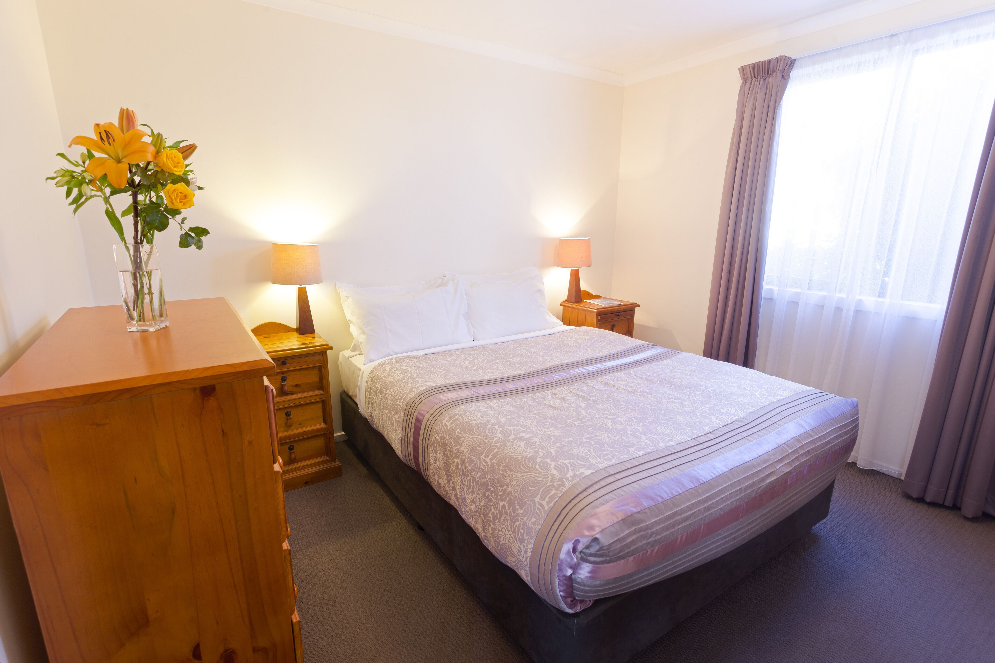 A hotel room with a bed, bedside tables, a lamp, a wooden dresser, and a vase with flowers.