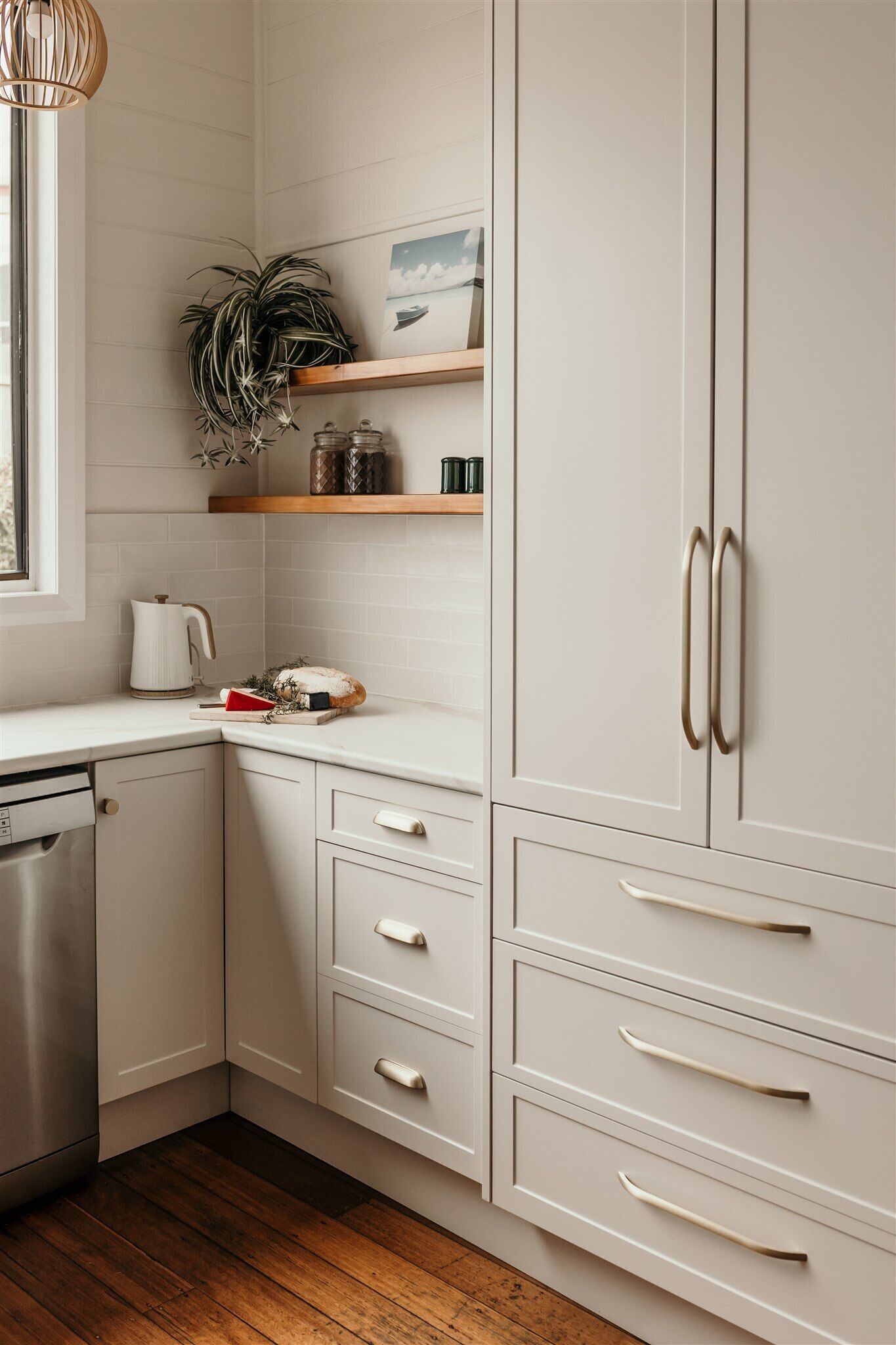 A kitchen with white cabinetry, wooden flooring, and a window with a plant on the counter.