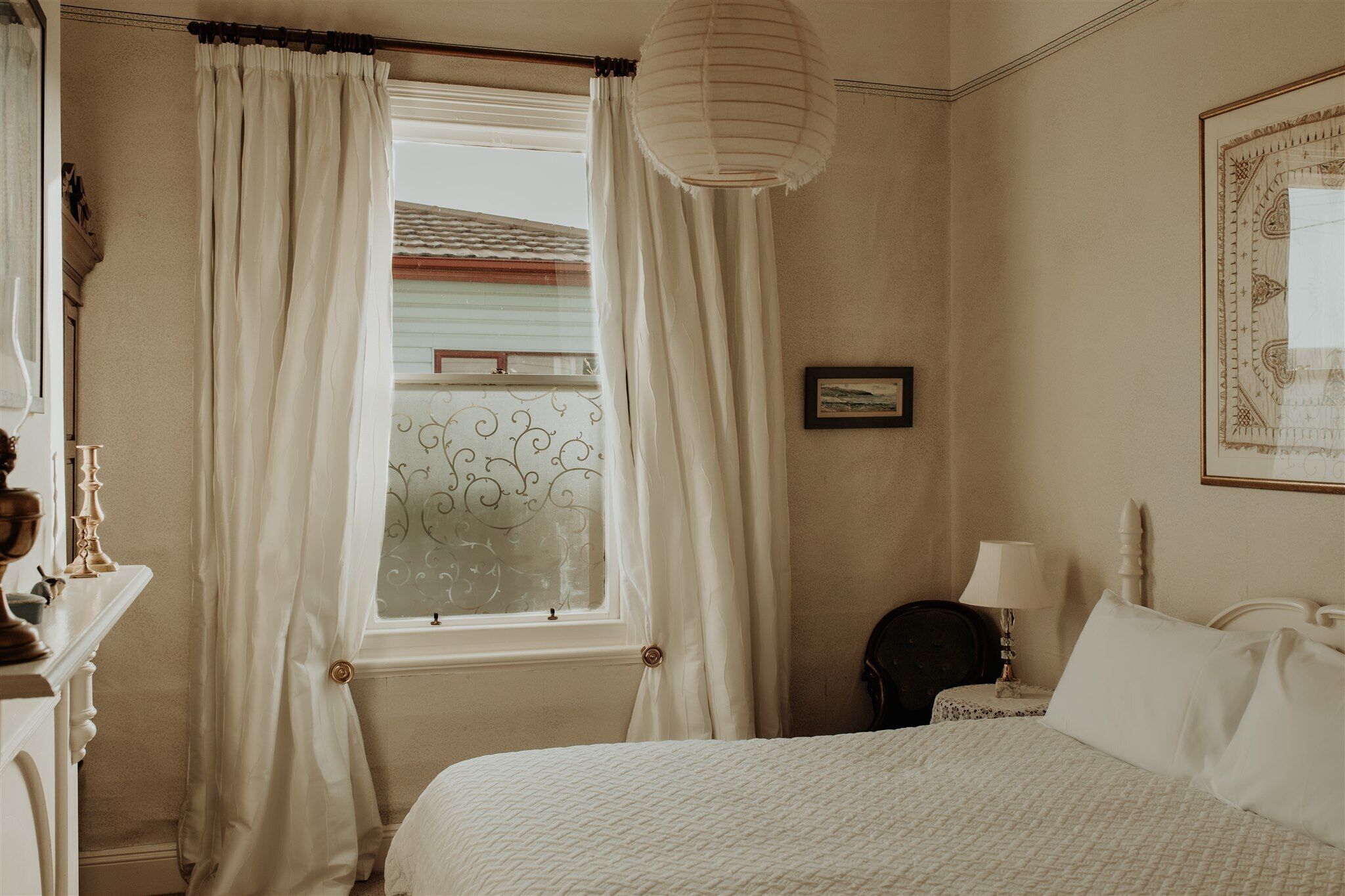 A bedroom with a bed, a nightstand with a lamp, a window with curtains, and a framed picture on the wall.