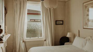 A bedroom with a bed, a nightstand with a lamp, a window with curtains, and a framed picture on the wall.