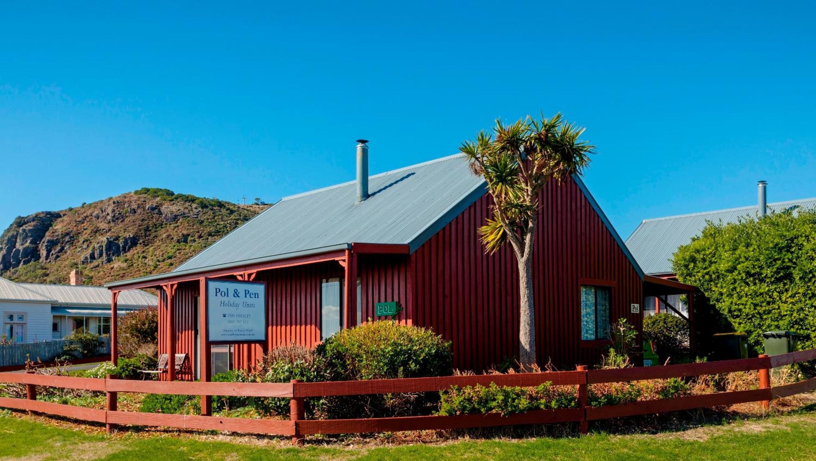 A red building with a sign for 'Pol & Pen Holiday Units' and a view of a mountainous landscape in the background.
