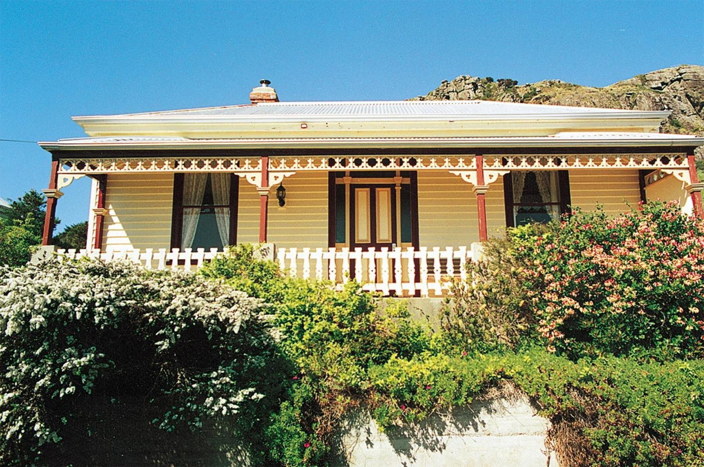 Una casa de dos pisos con valla de piquete blanca, un balcón y un muro de piedra al fondo.