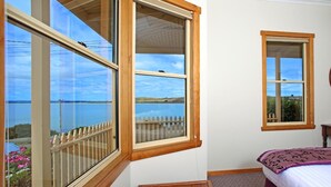 Un dormitorio con un ventanal que ofrece vistas a un lago y colinas, otra ventana más pequeña y un banco de madera.