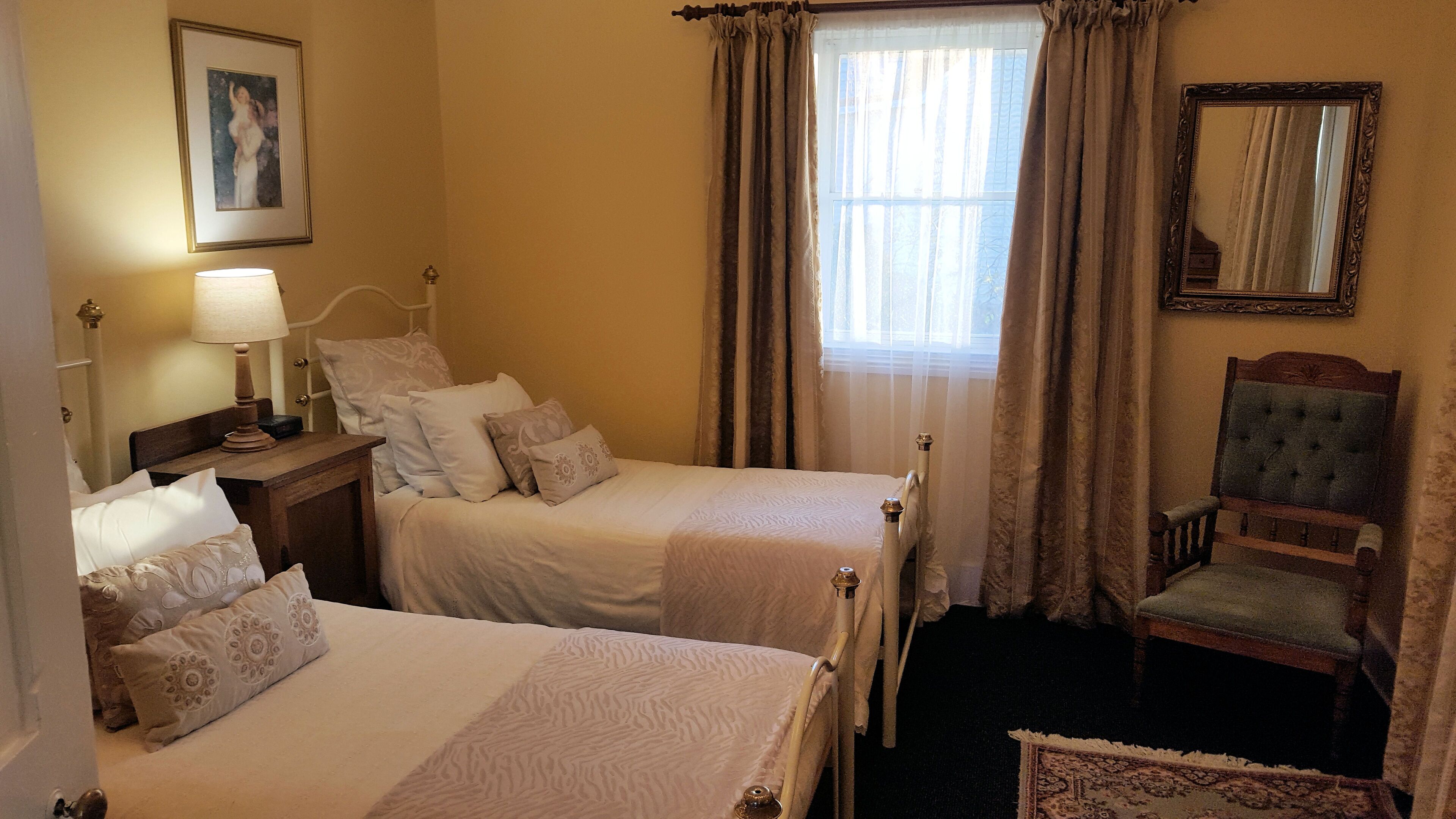 A hotel room with two beds, a chair, a bedside table with a lamp, a framed picture on the wall, and a window with curtains.