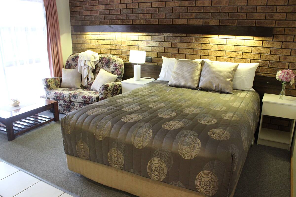 A hotel room with a large bed, a floral armchair, a nightstand with a lamp, and a brick wall.