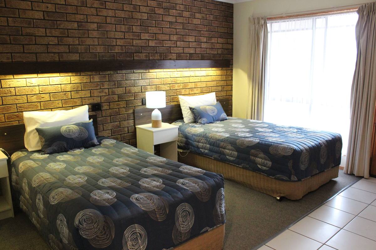 A hotel room with two beds, a brick wall, a nightstand with a lamp, and a window with curtains.
