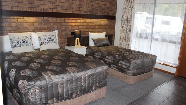 A double bed with a dark patterned bedspread, a nightstand with a lamp, and a window with sheer curtains. - Dunolly Golden Triangle Motel (Dunolly)
