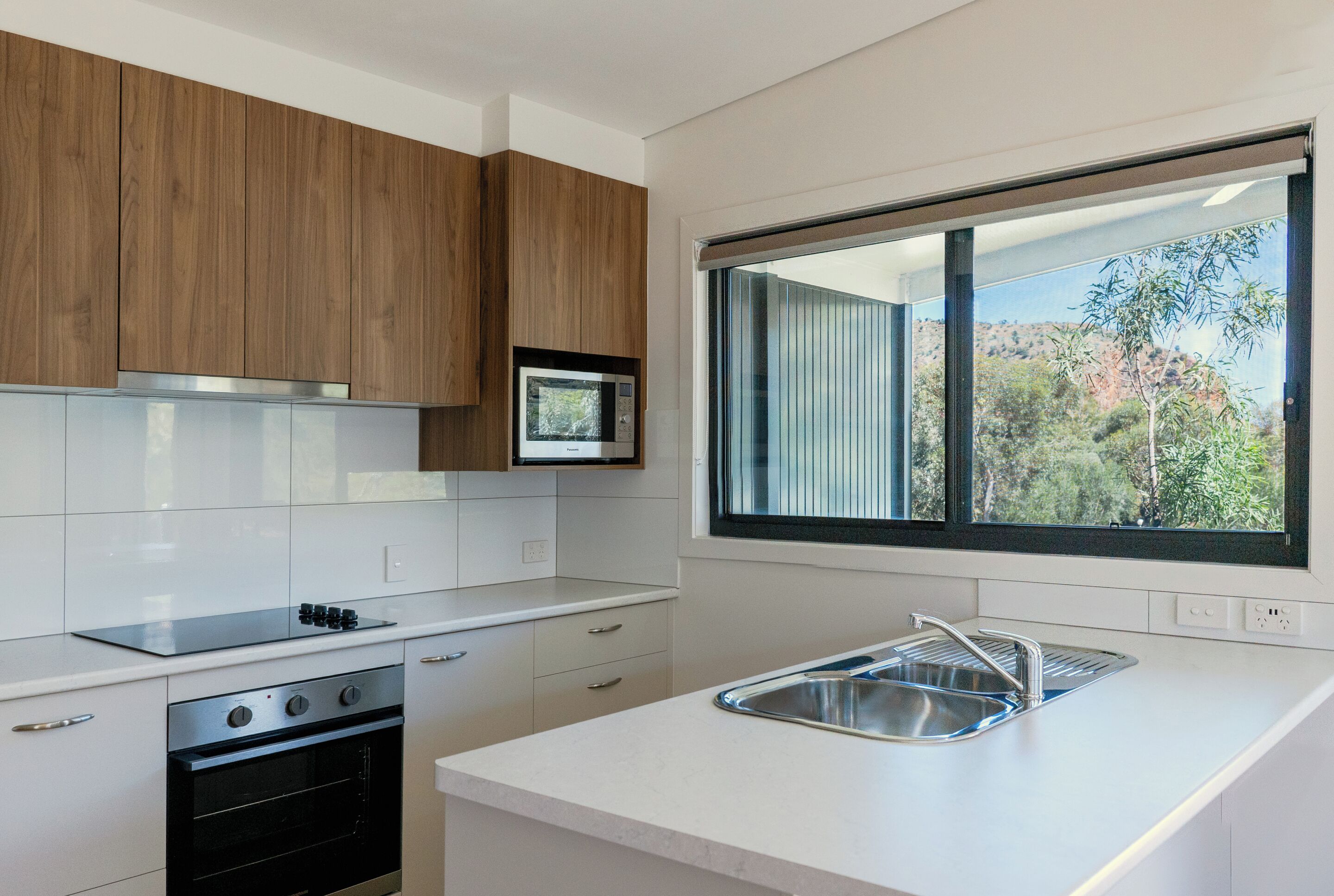 A modern kitchen with wooden cabinets, a built-in microwave, a stainless steel sink, and a large window offering a view of greenery and hills.