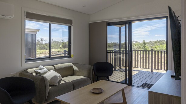 A modern living room with a sofa, a coffee table, and a TV. There is a balcony with a wooden deck and a view of the surrounding landscape. - Discovery Parks - Alice Springs (Ross)