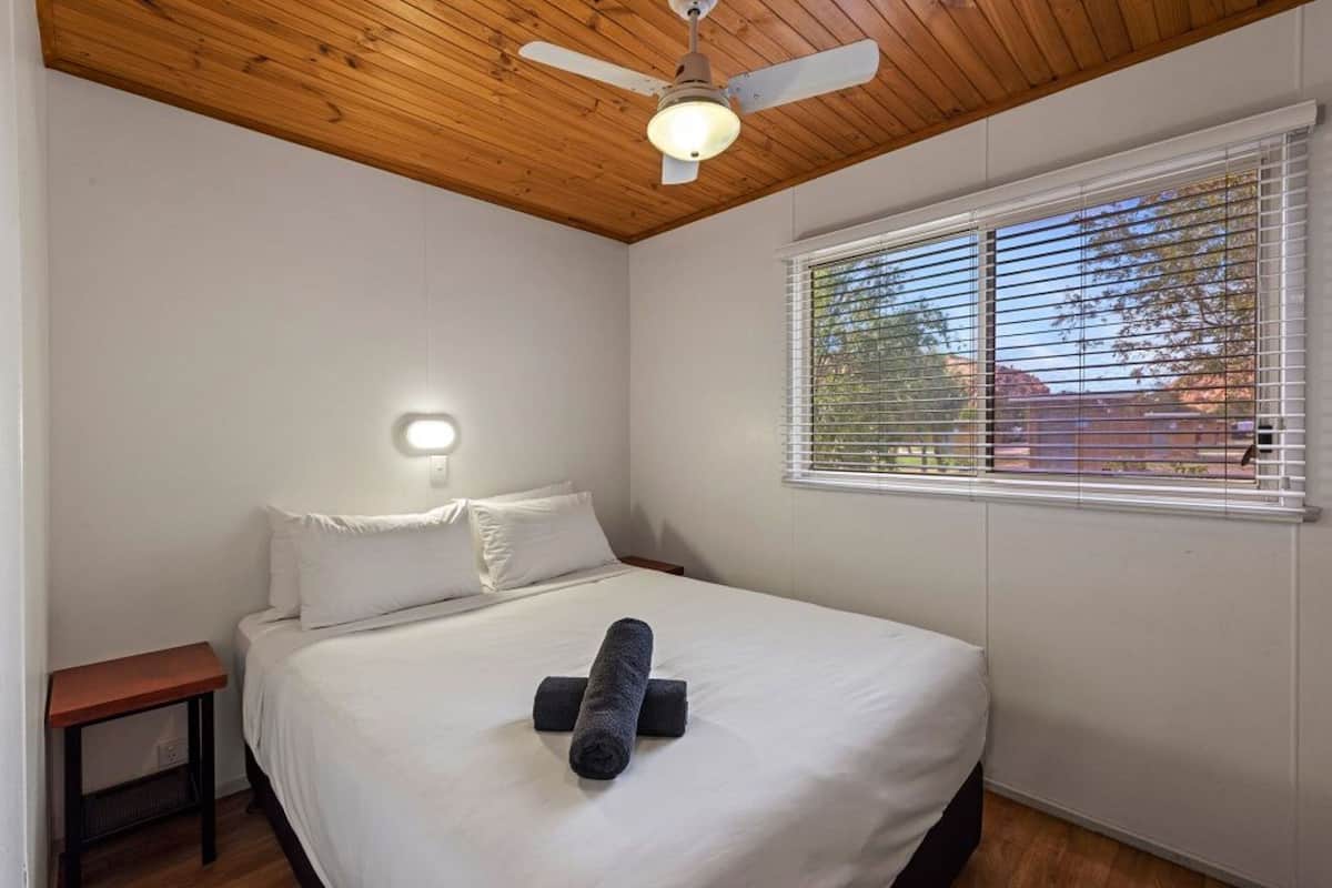 Quarto com teto de madeira, cama com roupa de cama branca, mesa de cabeceira e janela com persianas.