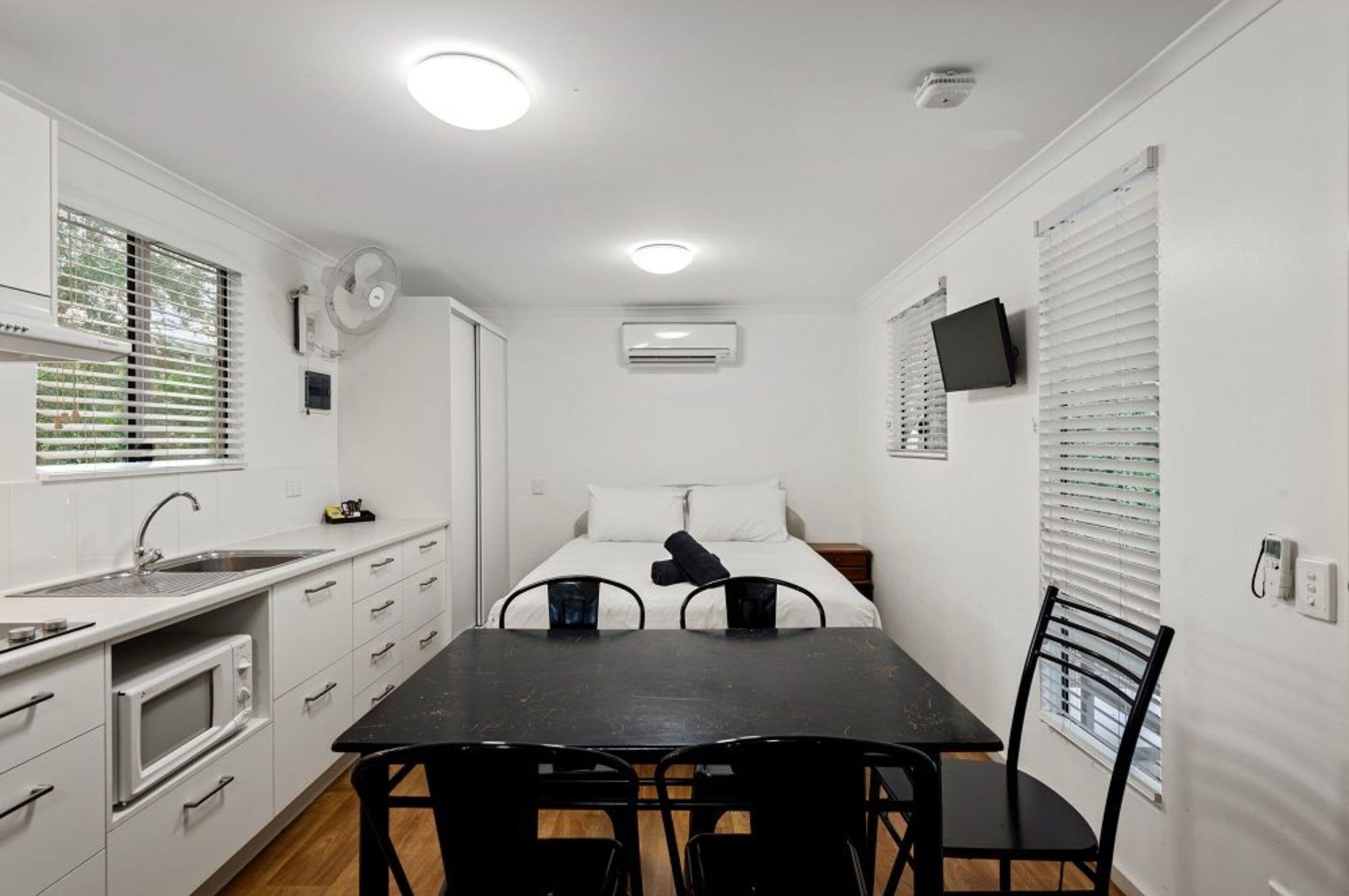 A compact room with a kitchenette, a bed, a TV, and a window with blinds.